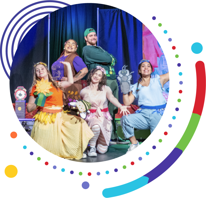 Five performers pose together on stage in colorful theatrical costumes, smiling and holding character puppets, framed by vibrant circular graphic elements.