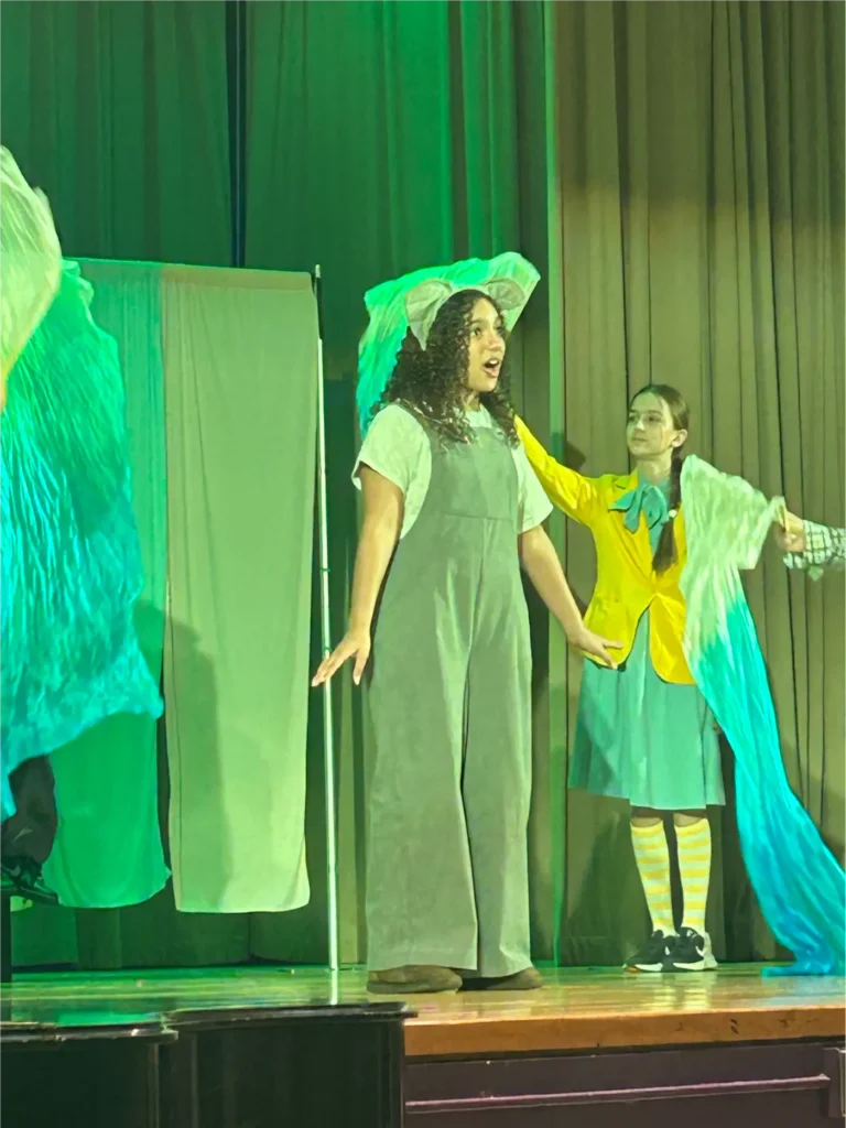 Young performers act on stage under green lighting, with a girl wearing mouse ears and a gray costume in the foreground and another performer in a yellow jacket and blue skirt holding flowing fabric behind her.