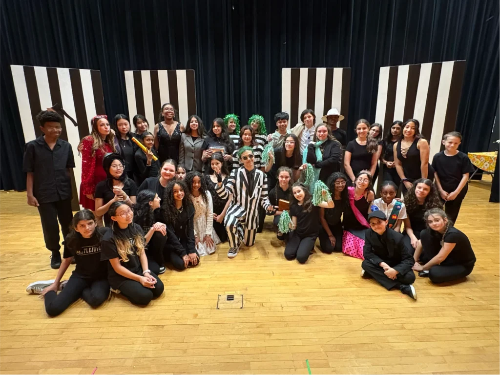Over 30 young performers smile together on stage, most dressed in black costumes, with others wearing red, white, and gray outfits, black and white striped suits, and green wigs.