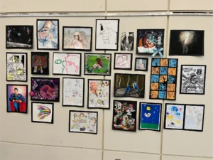 A gallery wall displays a diverse collection of framed student artwork, including colorful portraits, abstract illustrations, nature scenes, comic art, mixed media pieces, and imaginative drawings.