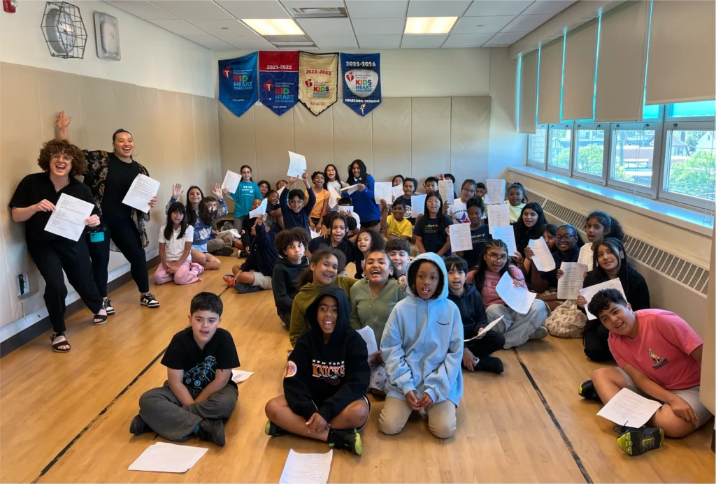 Approximately 40 young participants and two instructors smile together in a group photo, holding up their completed papers during a creative learning workshop.