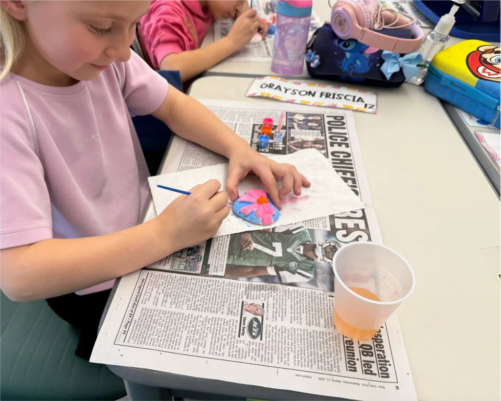 A smiling young girl looks down as she paints a heart shaped art project decorated with a bow, working over paper towels and newspaper with a cup of water nearby.