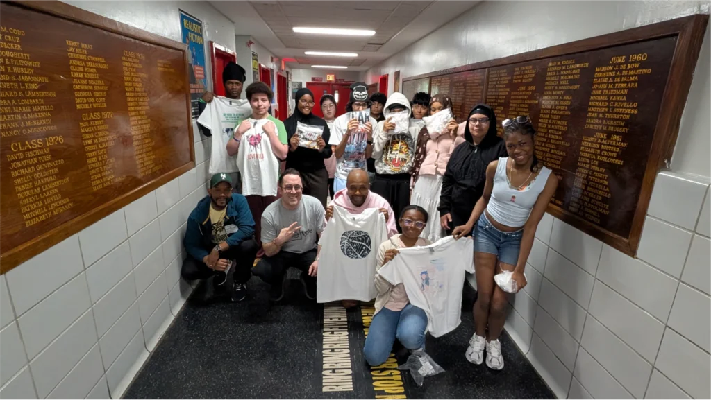 A group of approximately 15 teenagers smiles together in a hallway, proudly displaying their custom T-shirts featuring creative graphic designs made during an art workshop.