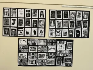 A collection of student artwork featuring black and white prints of animals, flowers, abstract patterns, letters, and creative illustrations arranged in three gallery displays on a classroom wall.