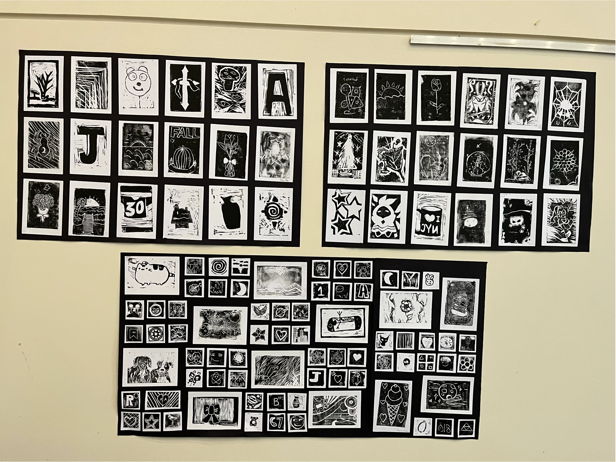 A collection of student artwork featuring black and white prints of animals, flowers, abstract patterns, letters, and creative illustrations arranged in three gallery displays on a classroom wall.
