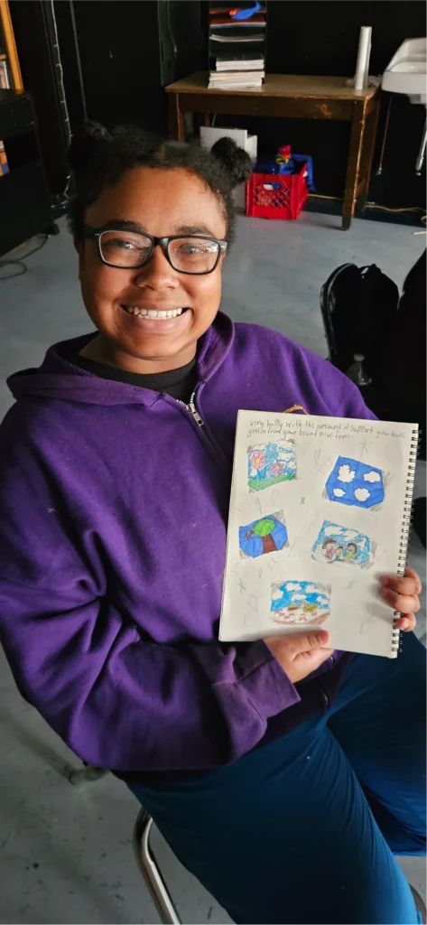 A smiling young artist in a purple hoodie displays a sketchbook featuring colorful drawings of nature, clouds, trees, and imaginative scenes during a creative art workshop.
