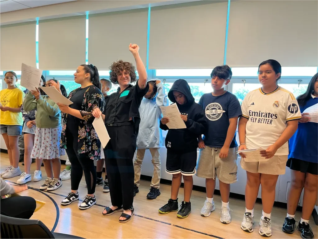 A theater instructor stands in the center with her arm raised and a smile, leading a group of young participants holding scripts during a collaborative theater workshop.