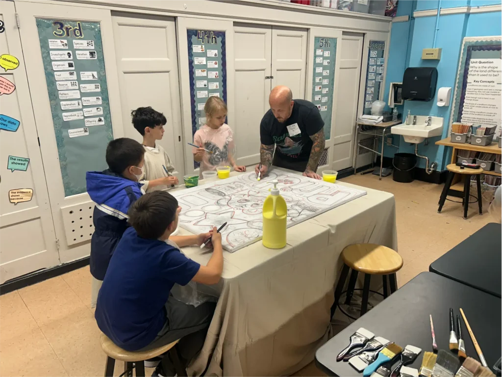 Participants work together on a large canvas during an art workshop, painting and outlining colorful illustrations with brushes and paint at a shared table.