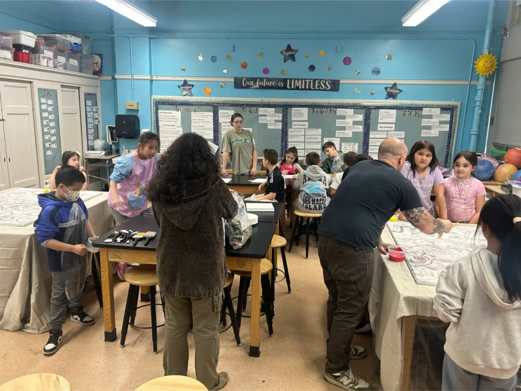 Group participants work together on a large canvas during an art workshop, painting and outlining colorful illustrations with brushes and paint at a shared table.