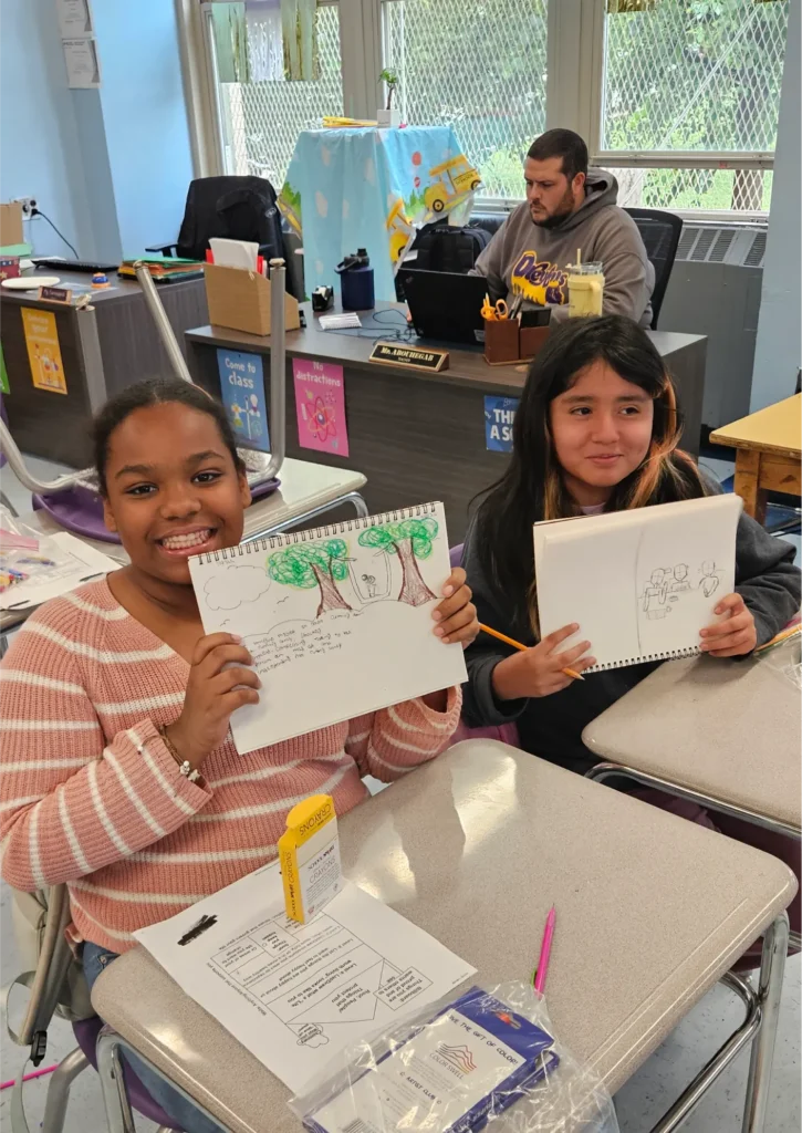Two students display their sketchbooks during an art workshop, sharing drawings of trees and other illustrations while seated at classroom desks.