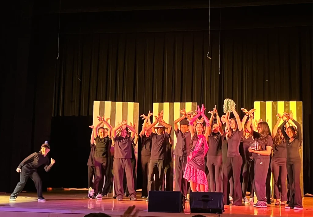 Young performers gather on stage under colorful lighting, with one performer striking a playful pose beside the group during a theater production or cast celebration.