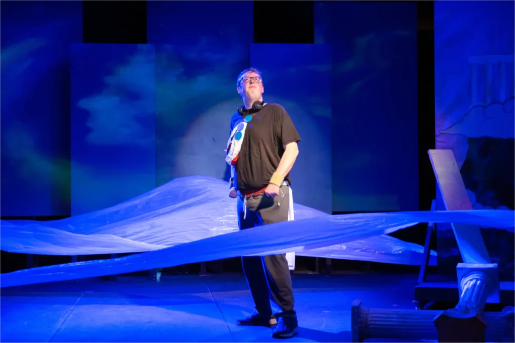 A theater production participant stands on stage surrounded by flowing blue fabric and scenic props under dramatic blue lighting during a stage production.