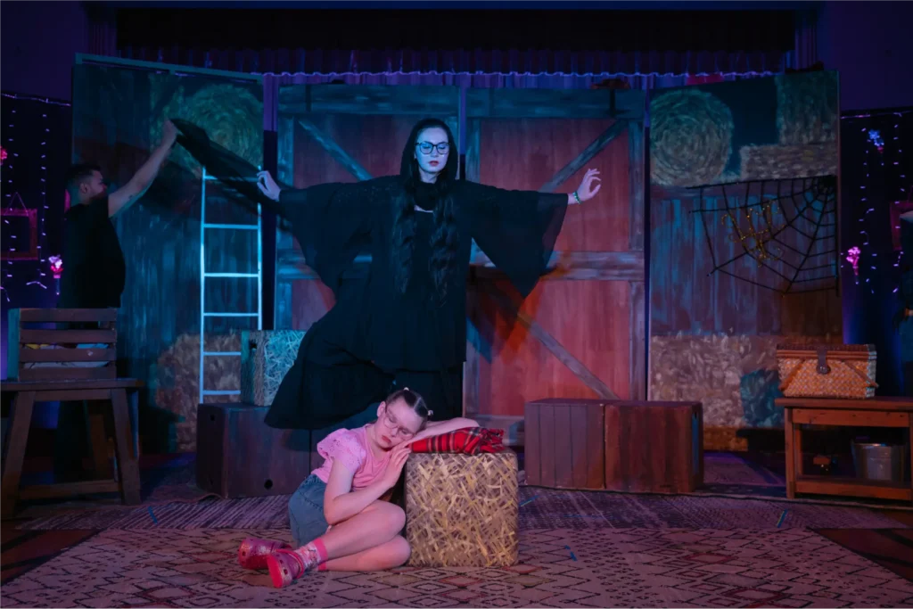 A performer in a flowing black costume stands with arms outstretched on a theatrical set, while a young girl rests against a haystack with her eyes closed. Blue and red stage lighting illuminates the rustic scenery.