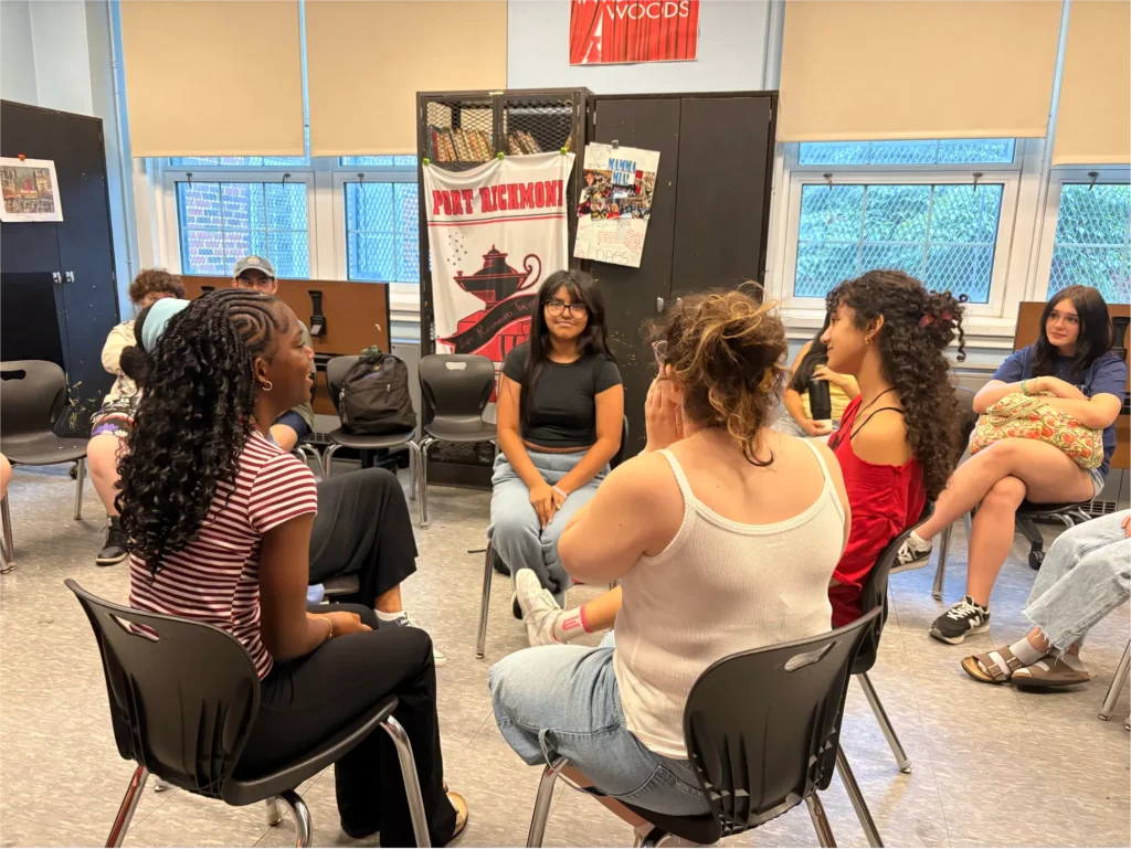 Young adults sit in a circle during a theater workshop, engaging in conversation and group activities in a creative learning environment.